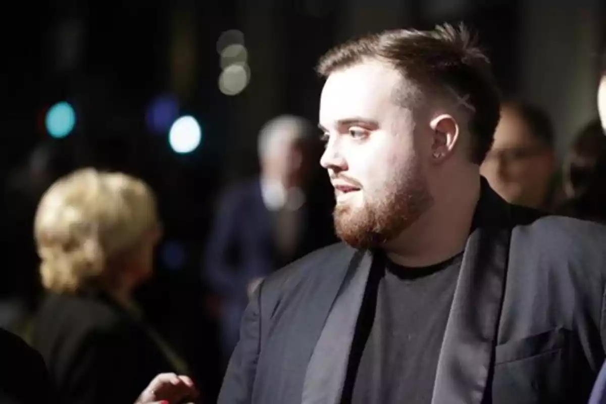 Hombre con barba y chaqueta oscura en un evento social con personas desenfocadas al fondo