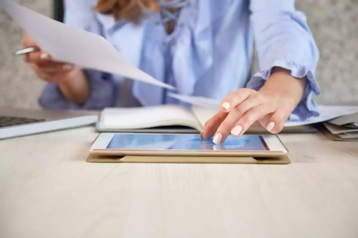 Persona trabajando en una mesa con una tablet, papeles y una computadora portátil
