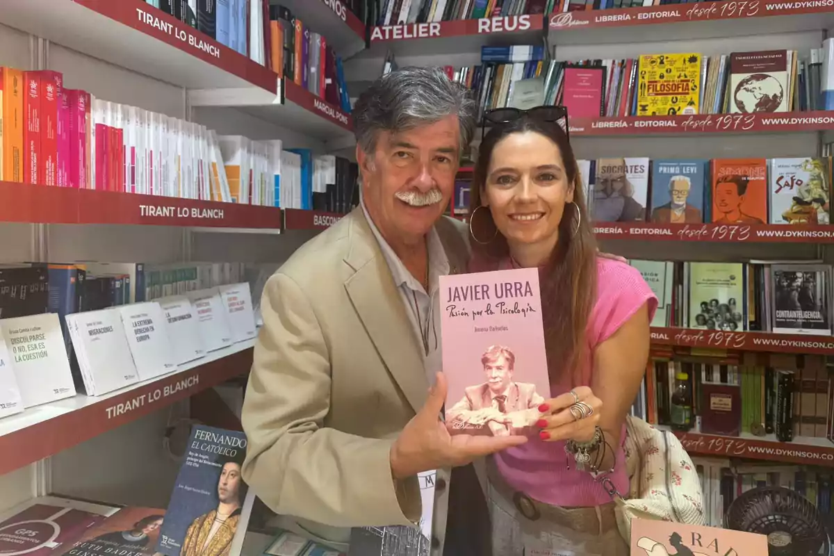 Un hombre y una mujer sonríen mientras sostienen un libro en una librería rodeados de estantes llenos de libros