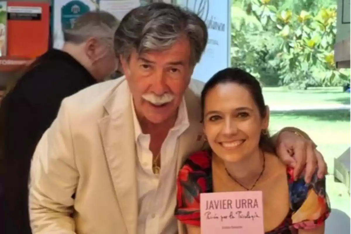 Un hombre de cabello canoso y bigote abraza a una mujer sonriente que sostiene un libro rosa con el nombre Javier Urra en la portada, ambos posan juntos en un entorno luminoso con vegetación al fondo.