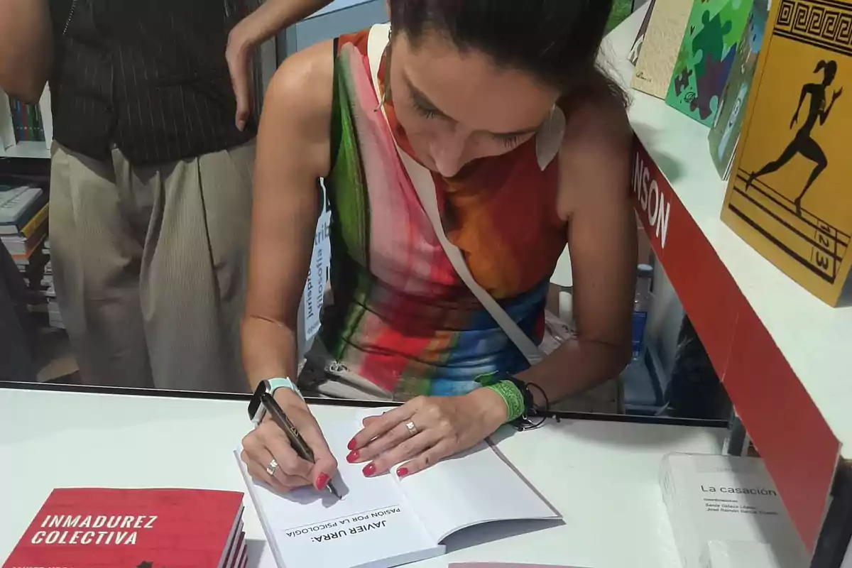 Mujer firmando un libro en una mesa durante una feria de libros, con varios ejemplares y personas alrededor