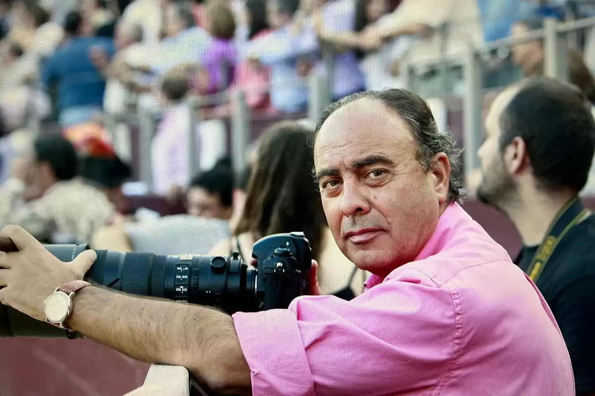 Hombre con camisa rosa sosteniendo una cámara profesional en un evento al aire libre con gente al fondo