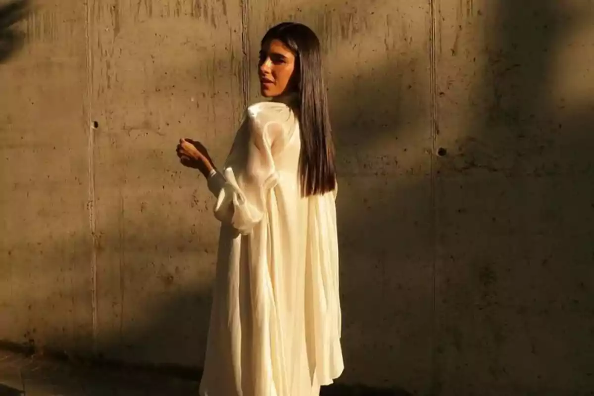 Mujer de cabello largo y liso vestida con una prenda blanca suelta de manga larga posando de perfil frente a una pared de concreto iluminada por la luz del sol Mujer de cabello largo y liso vestida con una prenda blanca suelta de manga larga posando de perfil frente a una pared de concreto iluminada por la luz del sol