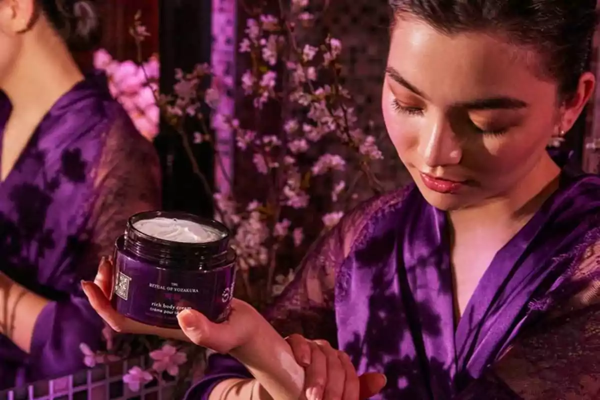 Mujer con bata morada aplicando crema corporal de un envase púrpura en un ambiente decorado con flores rosas Mujer con bata morada aplicando crema corporal de un envase púrpura en un ambiente decorado con flores rosas