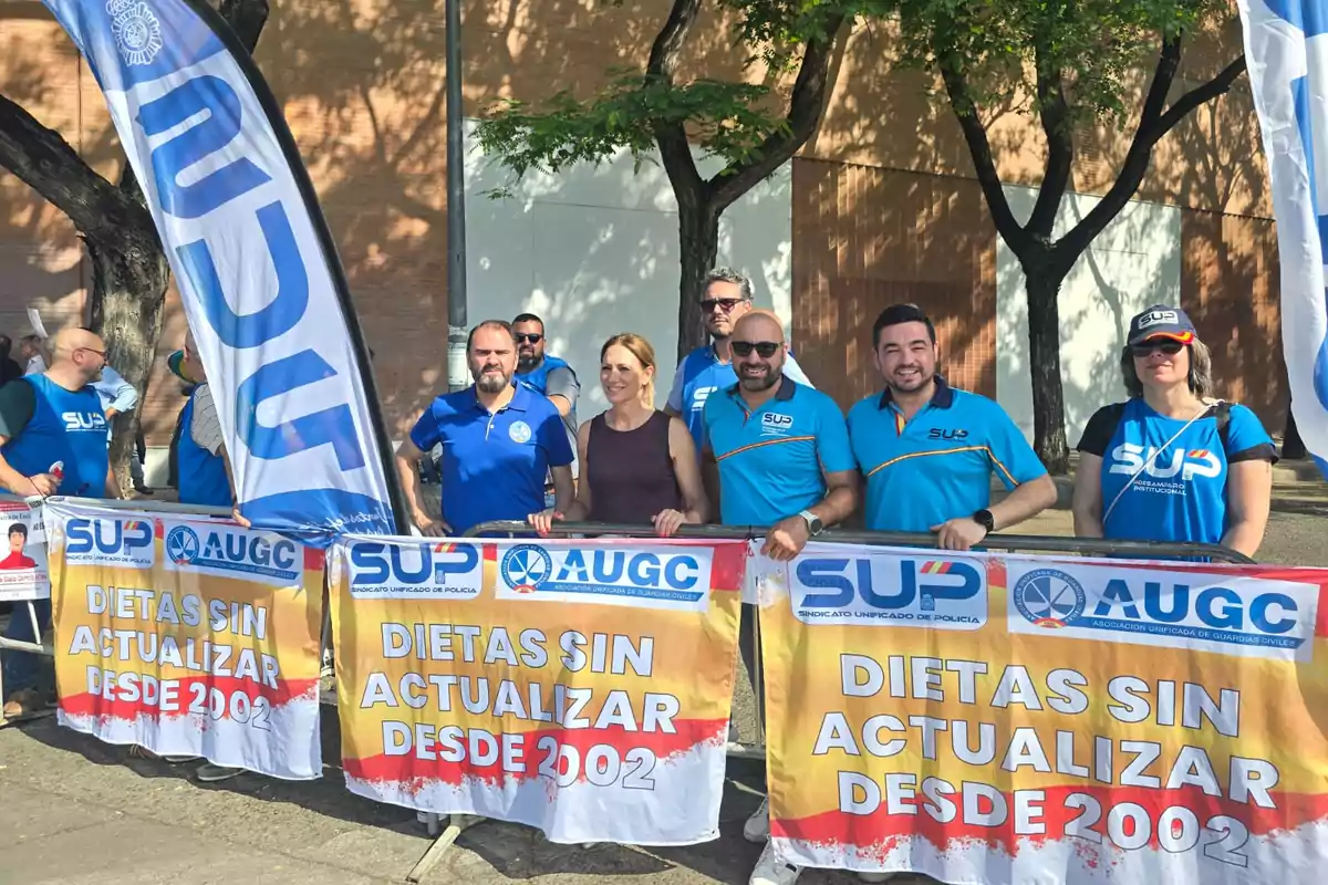 Un grupo de personas con camisetas azules sostiene pancartas que reclaman la actualización de dietas desde 2002 frente a una calle arbolada y soleada