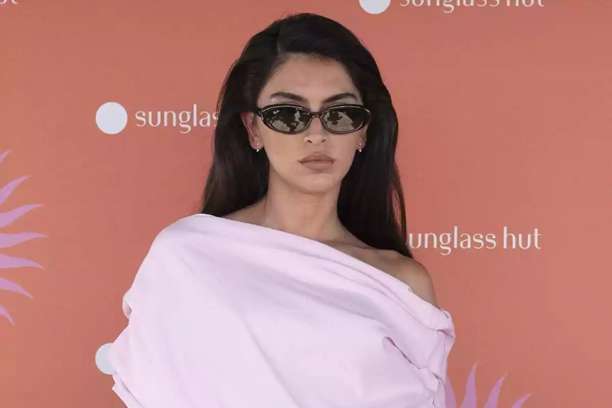 Lucía Rivera con cabello largo y oscuro usando gafas de sol y un vestido rosa claro posando frente a un fondo naranja con el logo de una tienda de gafas Lucía Rivera con cabello largo y oscuro usando gafas de sol y un vestido rosa claro posando frente a un fondo naranja con el logo de una tienda de gafas