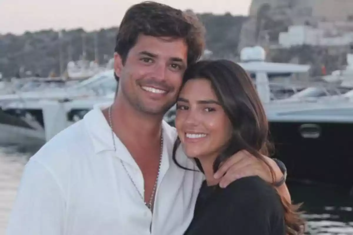 Pareja joven sonriendo y abrazándose frente a un puerto con yates al atardecer Pareja joven sonriendo y abrazándose frente a un puerto con yates al atardecer