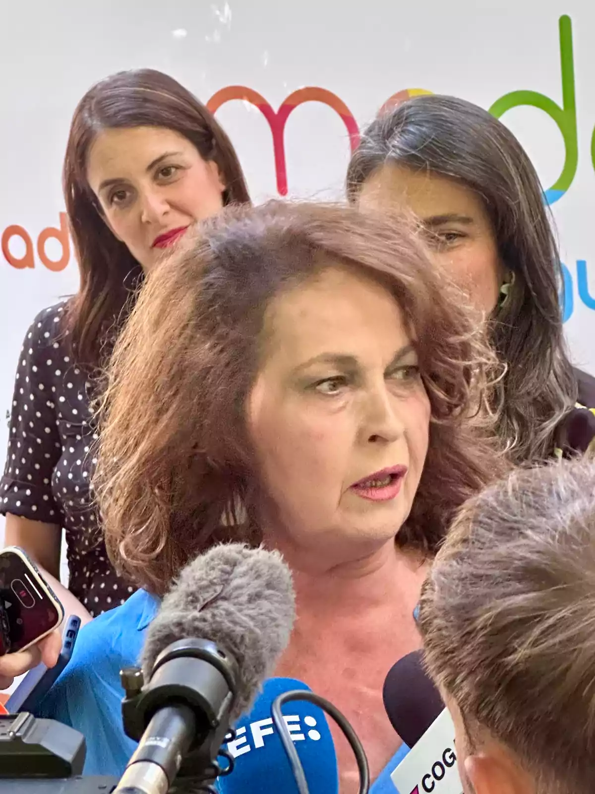 Mujer de cabello rizado hablando ante varios micrófonos rodeada de periodistas y con dos mujeres al fondo