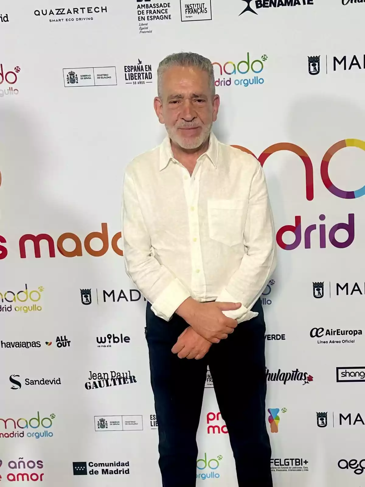 Hombre de cabello canoso y barba corta, vestido con camisa blanca y pantalón oscuro, posando frente a un fondo con logotipos de patrocinadores y el texto Madrid Orgullo
