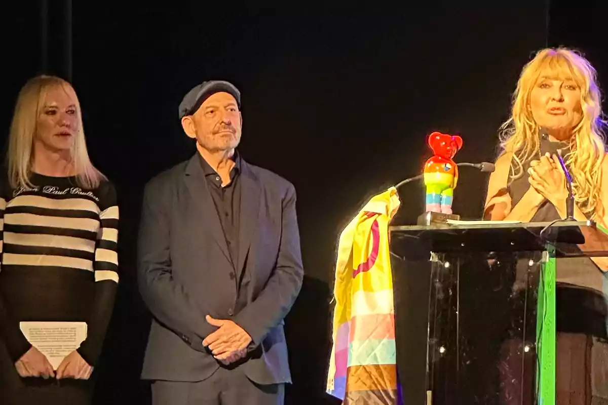 Tres personas en un escenario durante una ceremonia, una mujer habla en un podio decorado con una bandera de colores y una figura de oso con los colores del arcoíris, mientras las otras dos personas observan