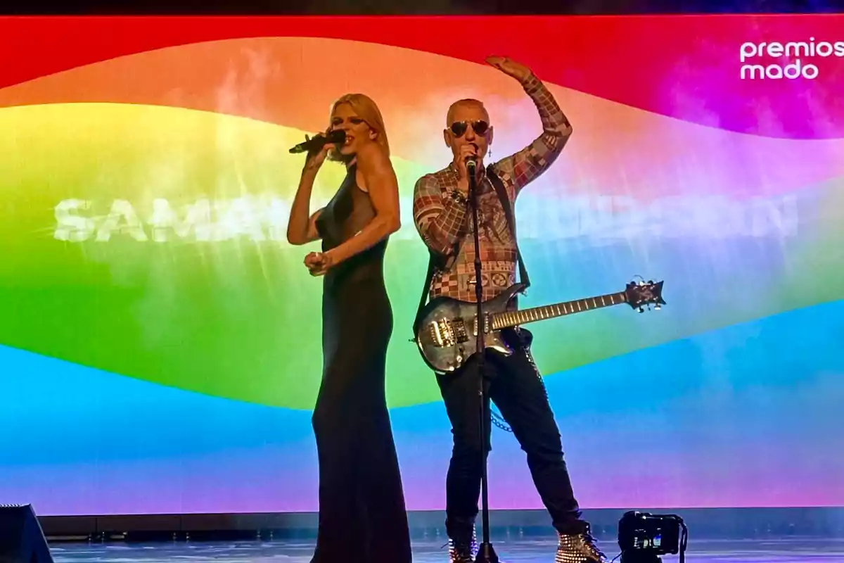 Dos personas cantan en el escenario frente a una pantalla con los colores del arcoíris y el texto premios mado, una de ellas sostiene una guitarra eléctrica.