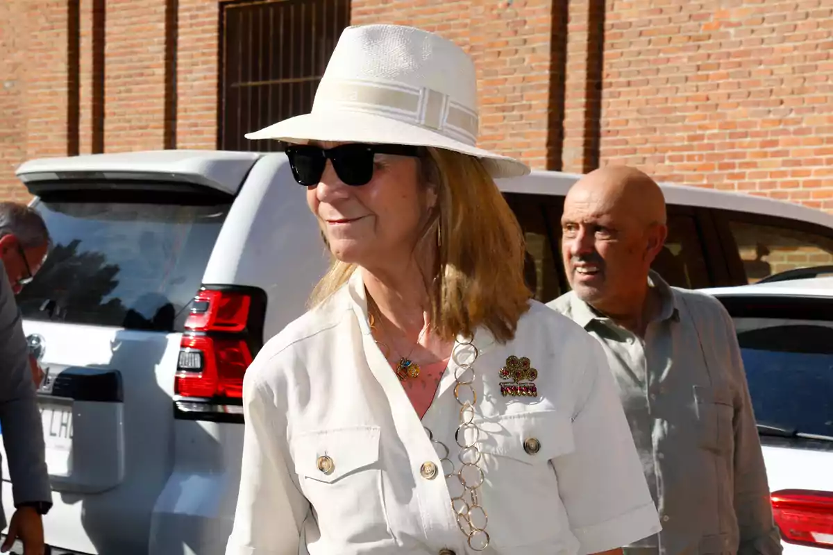 La Infanta Elena, en una imagen de archivo, con sombrero blanco y gafas de sol sonríe al aire libre junto a un hombre, ambos frente a un coche blanco y una pared de ladrillo. La Infanta Elena, en una imagen de archivo, con sombrero blanco y gafas de sol sonríe al aire libre junto a un hombre, ambos frente a un coche blanco y una pared de ladrillo.