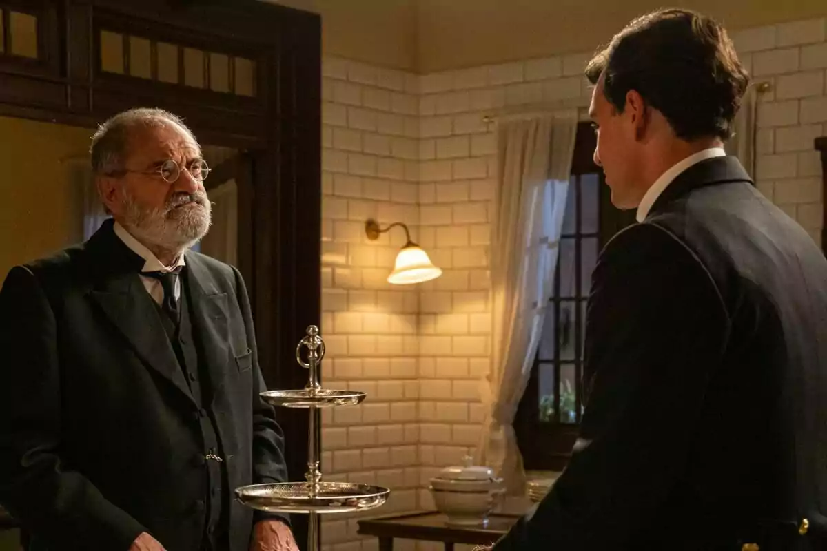 Joaquín Climent y Xavi Lock elegantemente vestidos conversan en una habitación con paredes de azulejos blancos y luz cálida en La Promesa, uno de ellos lleva gafas y barba canosa, mientras que el otro está de espaldas Joaquín Climent y Xavi Lock elegantemente vestidos conversan en una habitación con paredes de azulejos blancos y luz cálida en La Promesa, uno de ellos lleva gafas y barba canosa, mientras que el otro está de espaldas