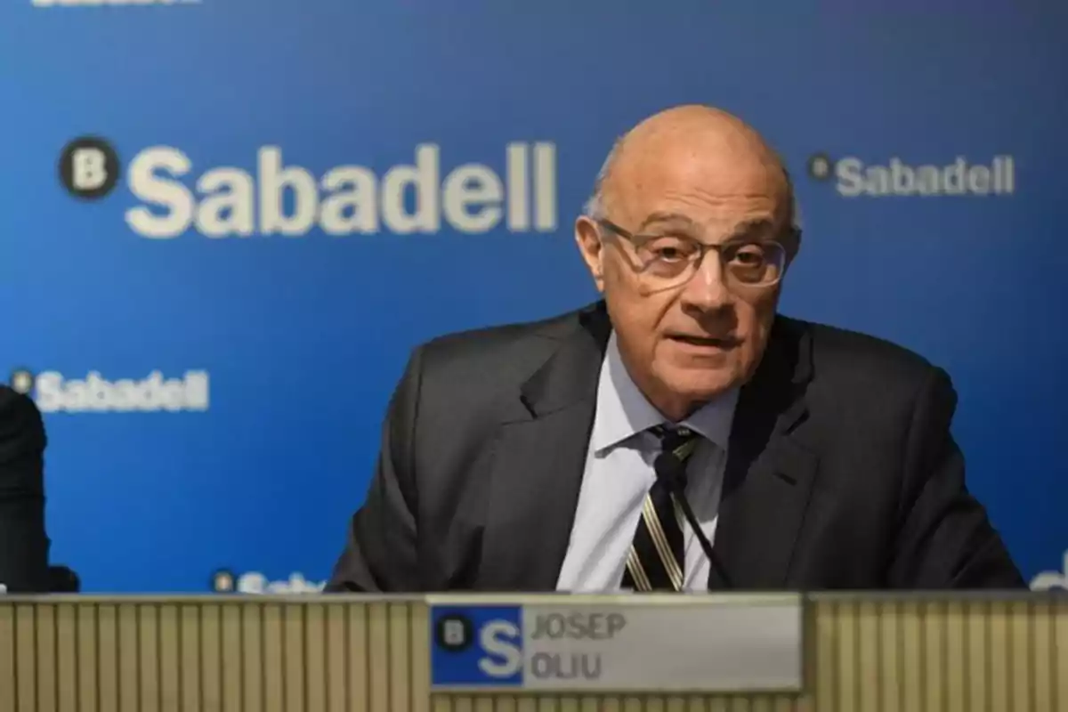 Hombre mayor con gafas y traje hablando en una conferencia frente a un fondo azul con el logo de Sabadell