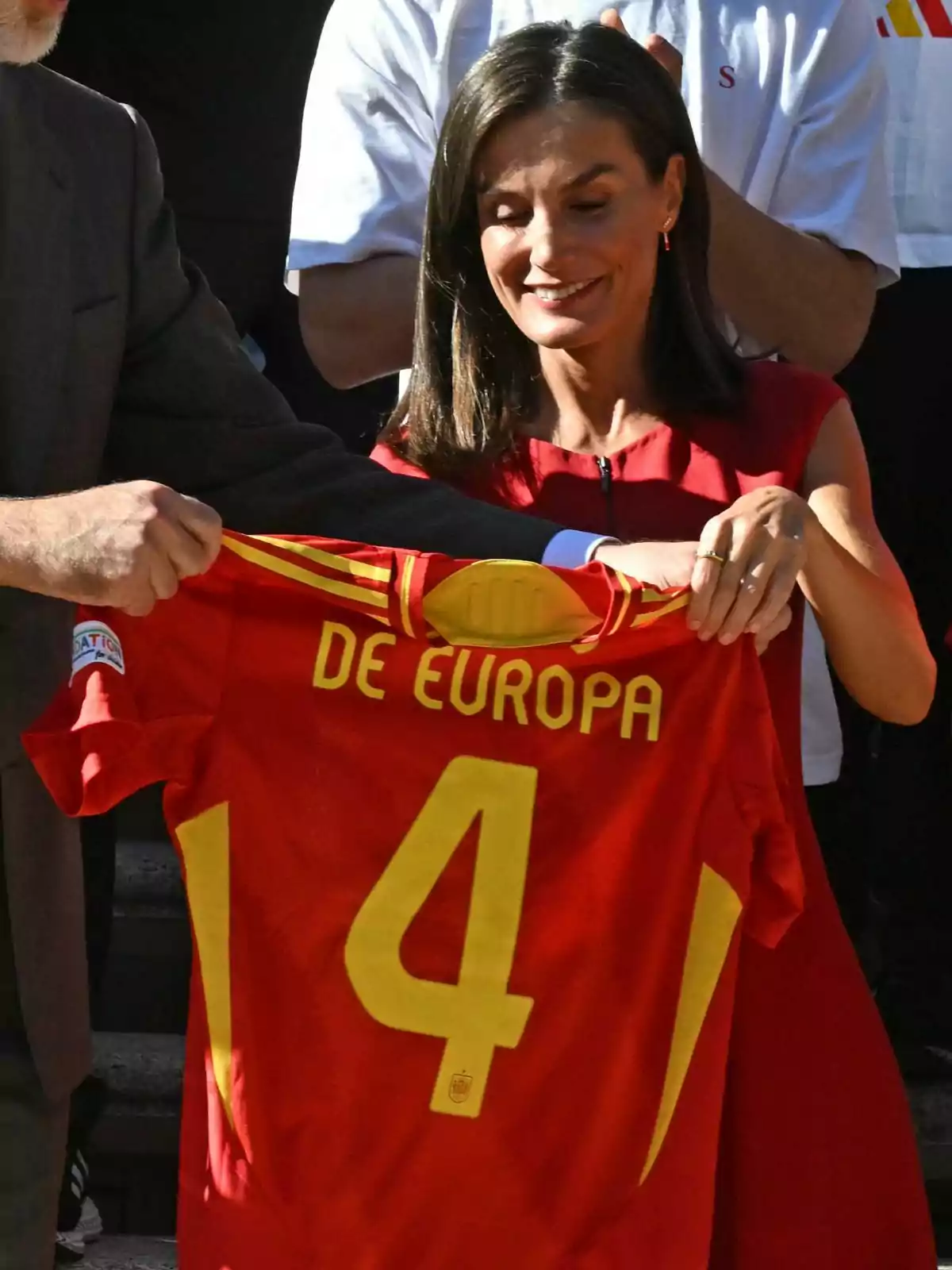 La reina, Letizia Ortiz, sonriente, sostiene una camiseta roja de la selección española de fútbol con el número 4 y la frase La reina, Letizia Ortiz, sonriente, sostiene una camiseta roja de la selección española de fútbol con el número 4 y la frase
