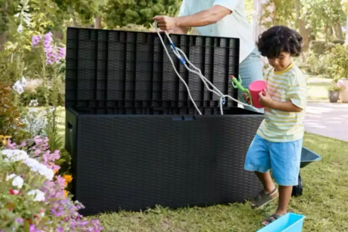 Un niño pequeño sostiene juguetes de jardín mientras un adulto abre un baúl negro grande en un jardín lleno de flores y vegetación Un niño pequeño sostiene juguetes de jardín mientras un adulto abre un baúl negro grande en un jardín lleno de flores y vegetación