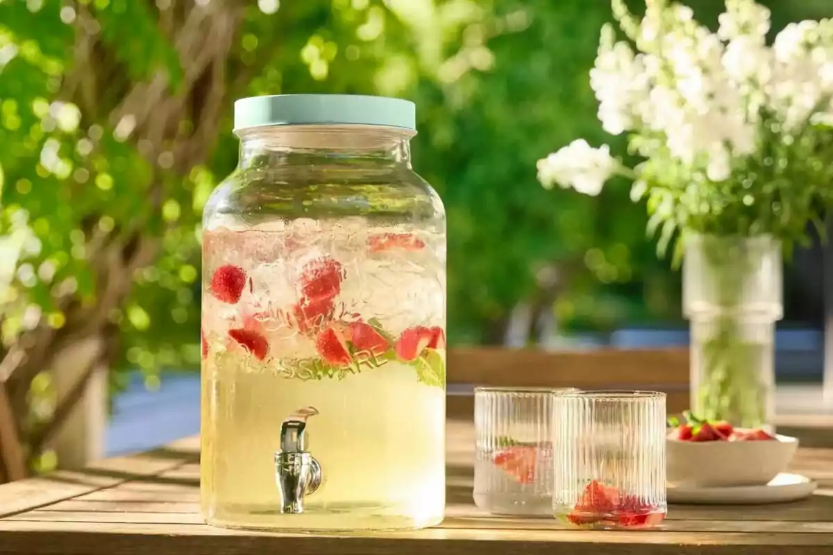 Jarra de vidrio con agua fresca de frutas y hielo sobre una mesa de madera al aire libre junto a dos vasos y un florero con flores blancas Jarra de vidrio con agua fresca de frutas y hielo sobre una mesa de madera al aire libre junto a dos vasos y un florero con flores blancas