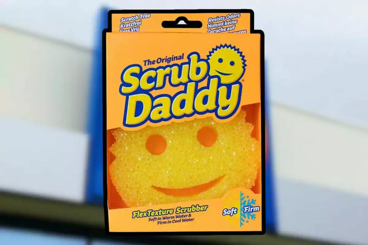 Esponja amarilla en forma de cara sonriente dentro de un empaque naranja con el nombre Scrub Daddy en letras grandes y azules Esponja amarilla en forma de cara sonriente dentro de un empaque naranja con el nombre Scrub Daddy en letras grandes y azules