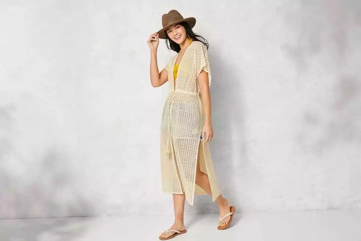 Mujer sonriente con sombrero de paja, vestido playero beige y sandalias posando frente a una pared clara Mujer sonriente con sombrero de paja, vestido playero beige y sandalias posando frente a una pared clara