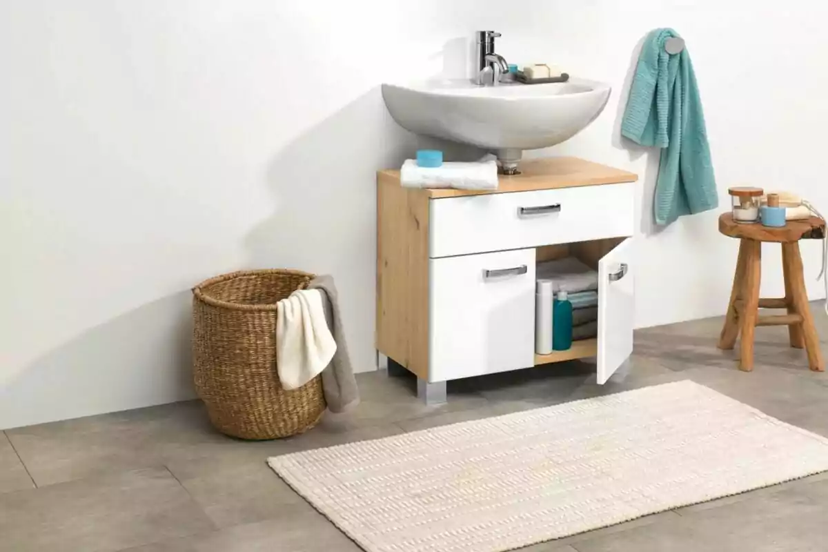 Baño moderno con lavabo blanco, mueble de madera, toallas, cesto de mimbre y alfombra clara sobre piso gris Baño moderno con lavabo blanco, mueble de madera, toallas, cesto de mimbre y alfombra clara sobre piso gris