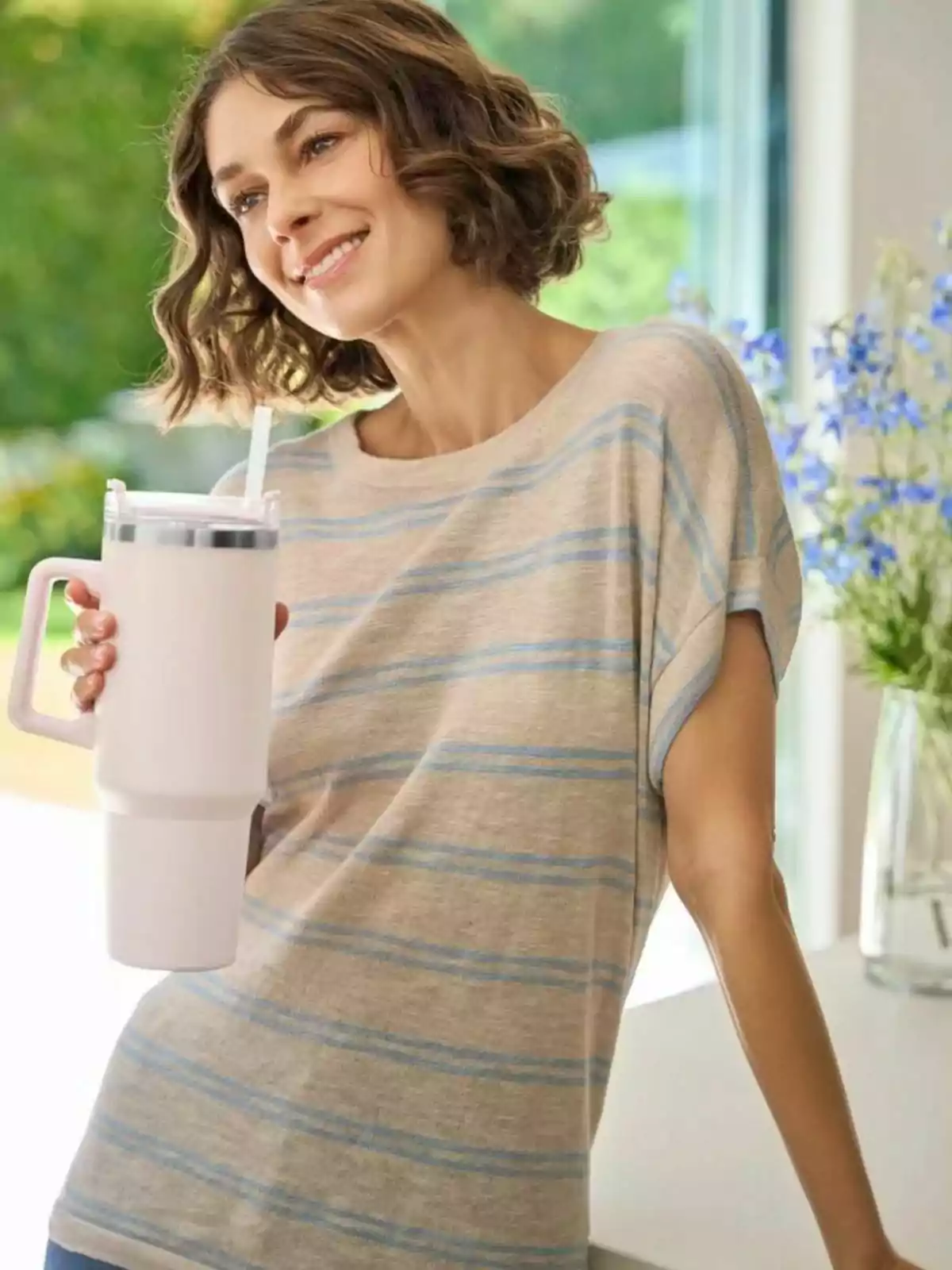 Mujer de cabello corto y ondulado sonriendo mientras sostiene un vaso grande con popote en una cocina iluminada y flores al fondo Mujer de cabello corto y ondulado sonriendo mientras sostiene un vaso grande con popote en una cocina iluminada y flores al fondo