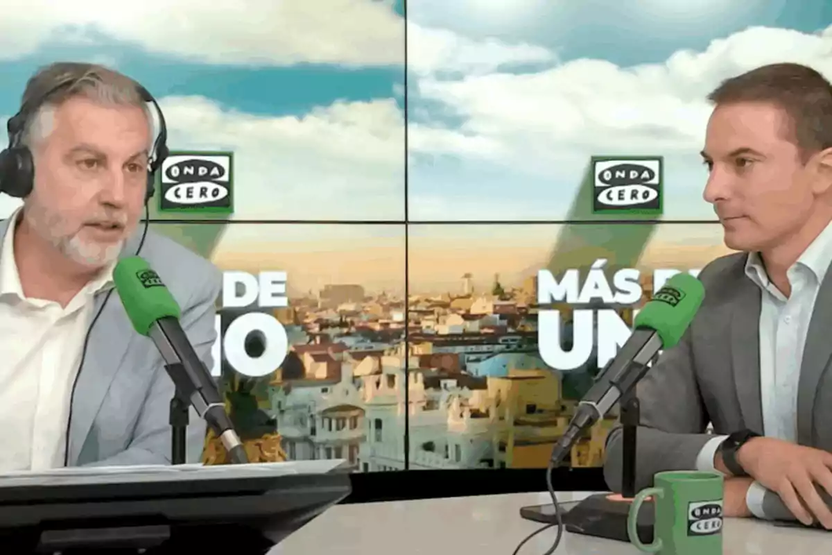 Dos hombres con traje conversan en un estudio de radio con micrófonos verdes y pantallas de fondo que muestran el logo de Onda Cero y la ciudad de Madrid Dos hombres con traje conversan en un estudio de radio con micrófonos verdes y pantallas de fondo que muestran el logo de Onda Cero y la ciudad de Madrid