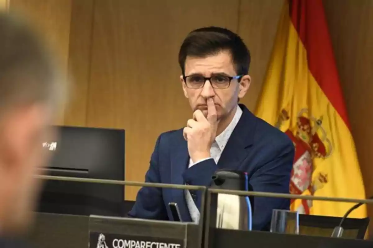 Hombre con gafas y traje azul sentado en una sala oficial con una bandera de España al fondo, parece pensativo y está apoyando la mano en su rostro. Hombre con gafas y traje azul sentado en una sala oficial con una bandera de España al fondo, parece pensativo y está apoyando la mano en su rostro.