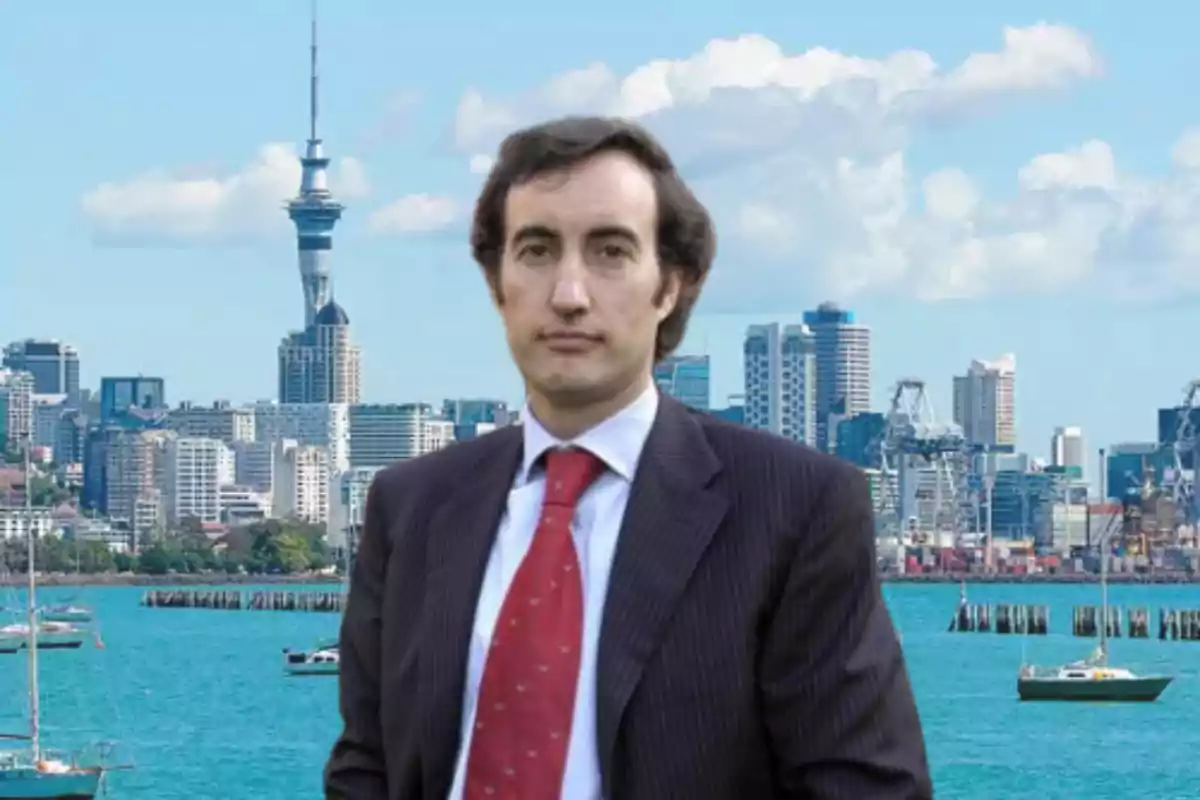 Hombre con traje y corbata roja posando frente a un skyline moderno con edificios altos y un cuerpo de agua con barcos