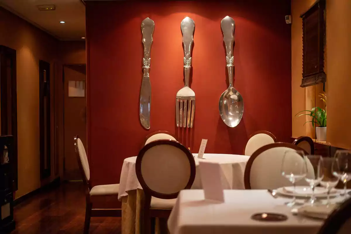Restaurante elegante con mesas redondas, sillas tapizadas y una pared decorada con cubiertos gigantes de metal Restaurante elegante con mesas redondas, sillas tapizadas y una pared decorada con cubiertos gigantes de metal