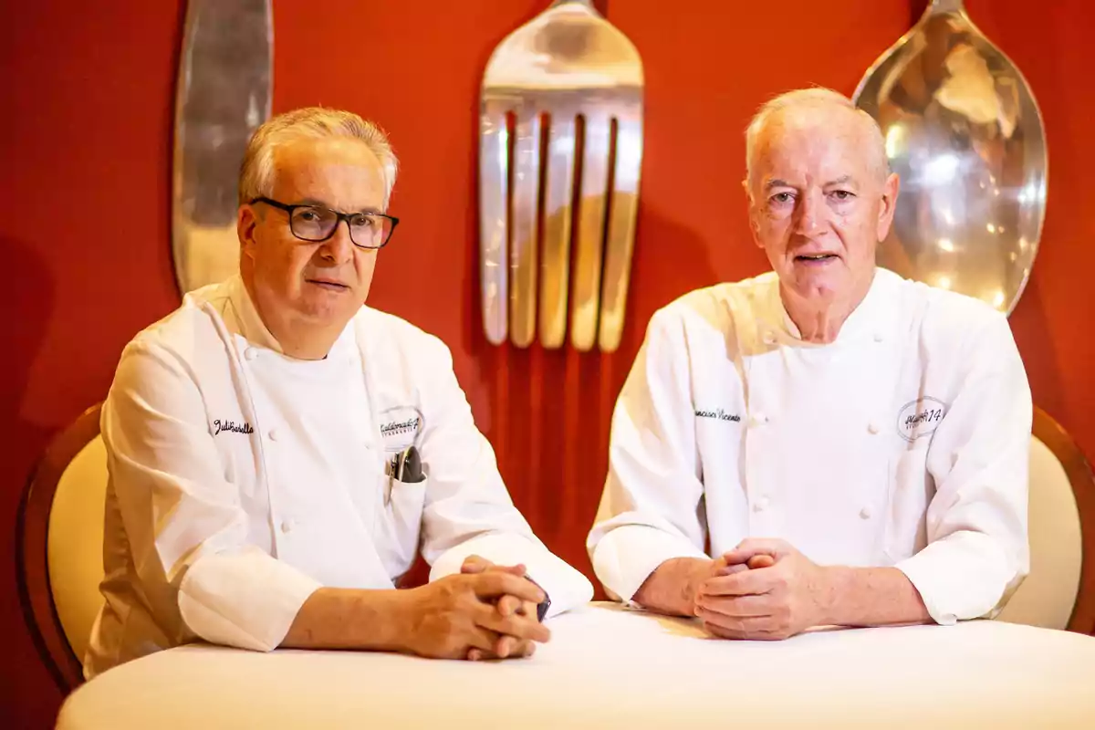 Dos chefs con uniforme blanco sentados en una mesa frente a una pared roja decorada con cubiertos gigantes