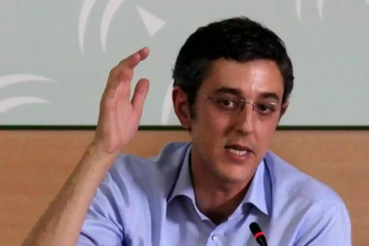 Hombre con gafas y camisa azul hablando frente a un micrófono y levantando una mano Hombre con gafas y camisa azul hablando frente a un micrófono y levantando una mano