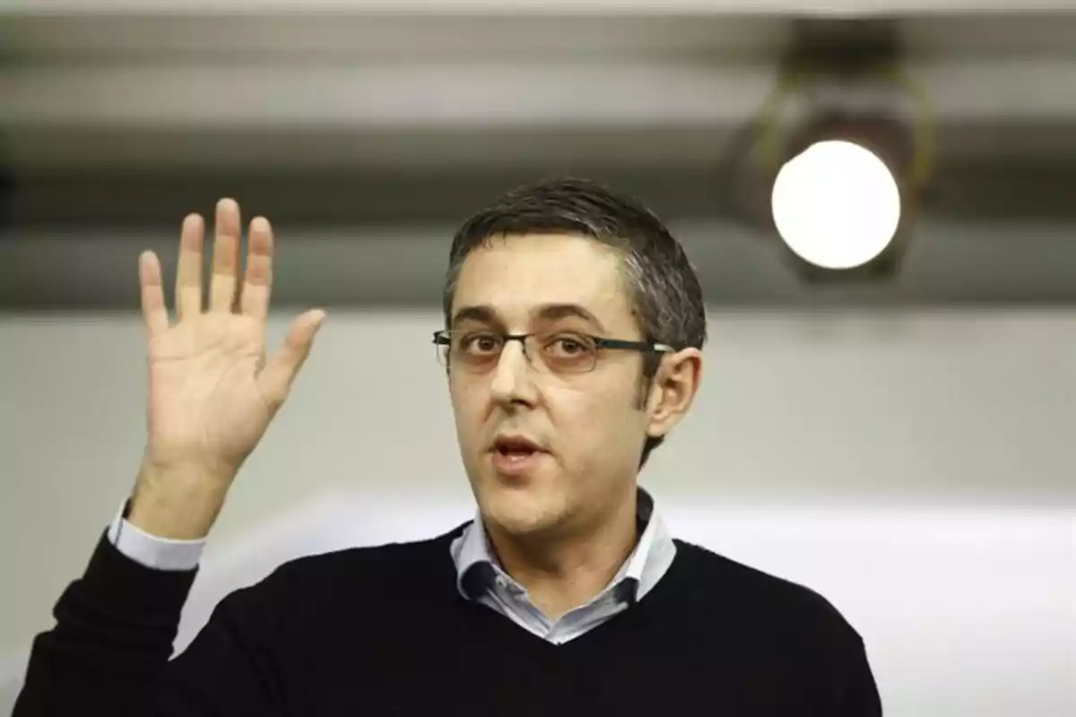 Hombre con gafas levantando la mano y hablando en un ambiente interior