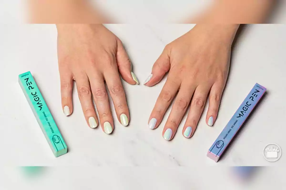Manos con uñas pintadas en tonos pastel iridiscentes de Mercadona sobre una superficie blanca junto a dos cajas de esmalte en forma de pluma. Manos con uñas pintadas en tonos pastel iridiscentes de Mercadona sobre una superficie blanca junto a dos cajas de esmalte en forma de pluma.