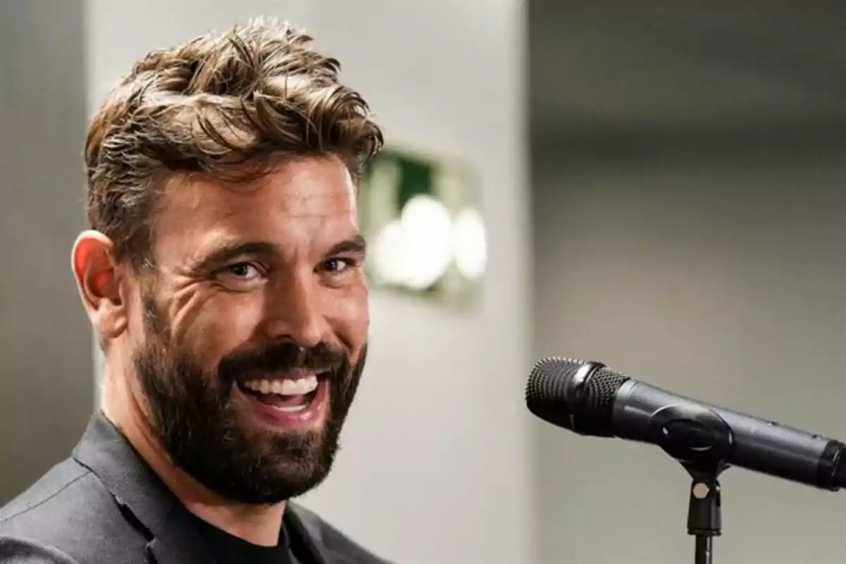 Hombre sonriente con barba frente a un micrófono en un ambiente interior