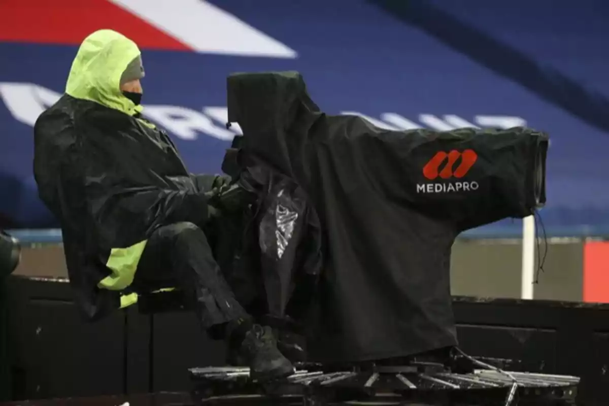 Persona con impermeable operando una cámara profesional cubierta con protección de lluvia en un evento deportivo Persona con impermeable operando una cámara profesional cubierta con protección de lluvia en un evento deportivo