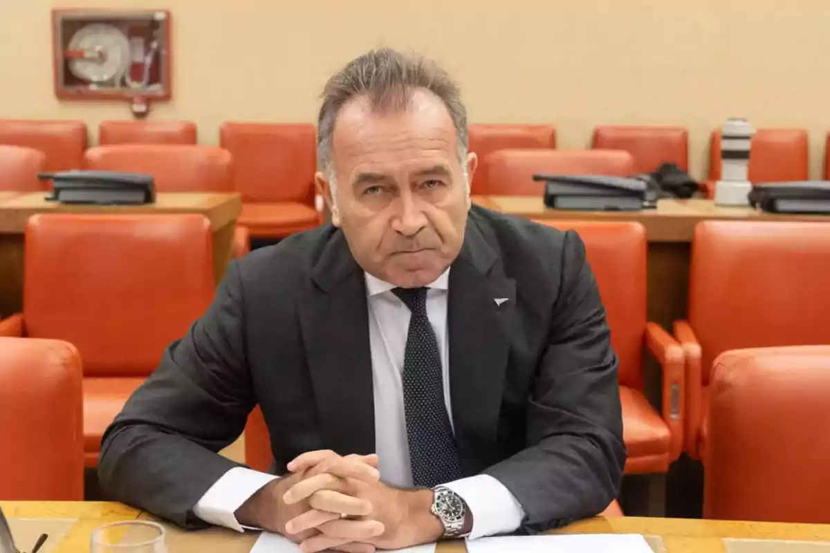 Hombre de traje oscuro sentado en una sala con sillas naranjas y expresión seria Hombre de traje oscuro sentado en una sala con sillas naranjas y expresión seria