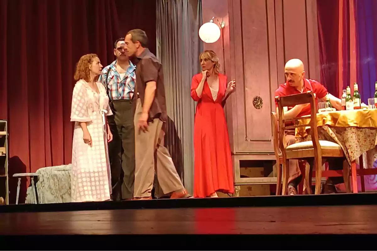 Cinco actores en el escenario durante una obra de teatro, tres de pie en el centro con expresiones intensas, una mujer al fondo con vestido rojo parece sorprendida y un hombre calvo está sentado a la mesa con botellas y vasos. Cinco actores en el escenario durante una obra de teatro, tres de pie en el centro con expresiones intensas, una mujer al fondo con vestido rojo parece sorprendida y un hombre calvo está sentado a la mesa con botellas y vasos.