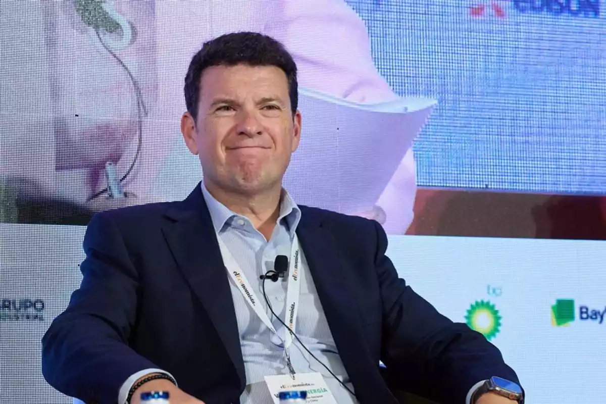 Hombre de cabello corto y oscuro con expresión sonriente sentado en un panel de conferencia usando saco azul y camisa clara con gafete y micrófono Hombre de cabello corto y oscuro con expresión sonriente sentado en un panel de conferencia usando saco azul y camisa clara con gafete y micrófono