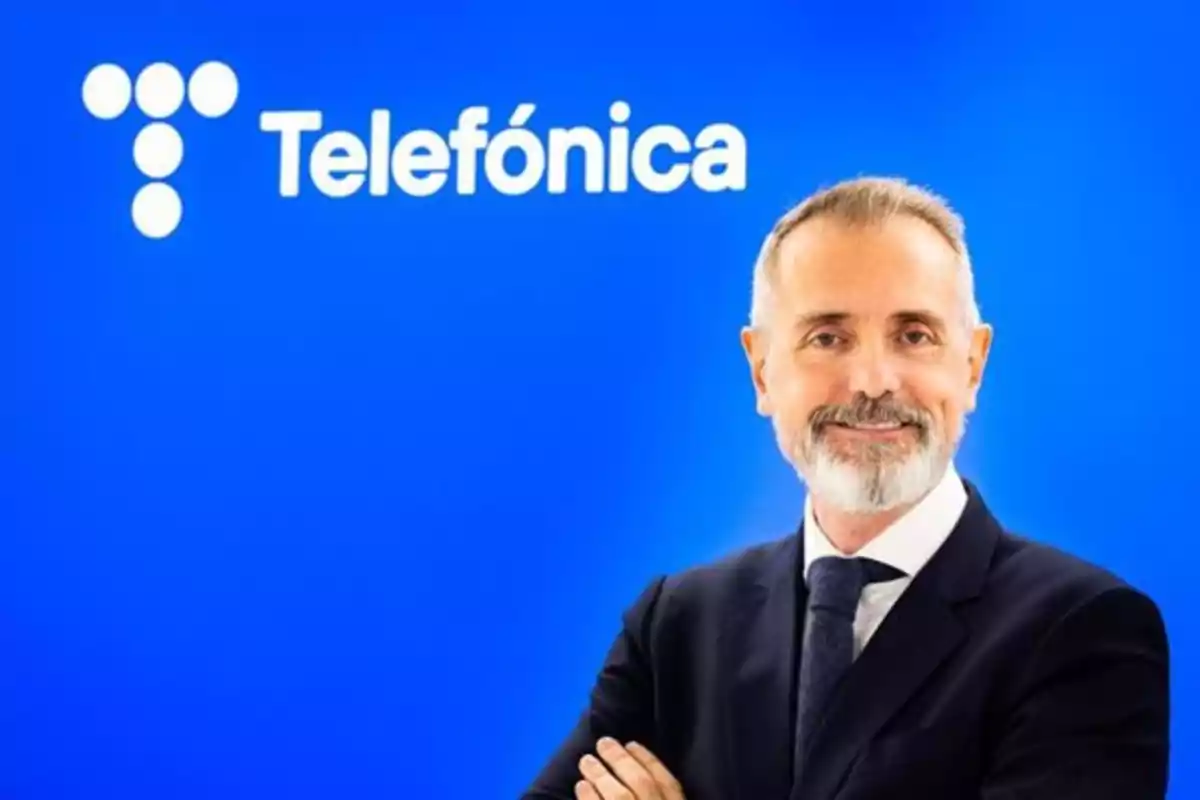 Hombre de traje con barba y cabello canoso sonríe frente a un fondo azul con el logo y nombre de Telefónica Hombre de traje con barba y cabello canoso sonríe frente a un fondo azul con el logo y nombre de Telefónica