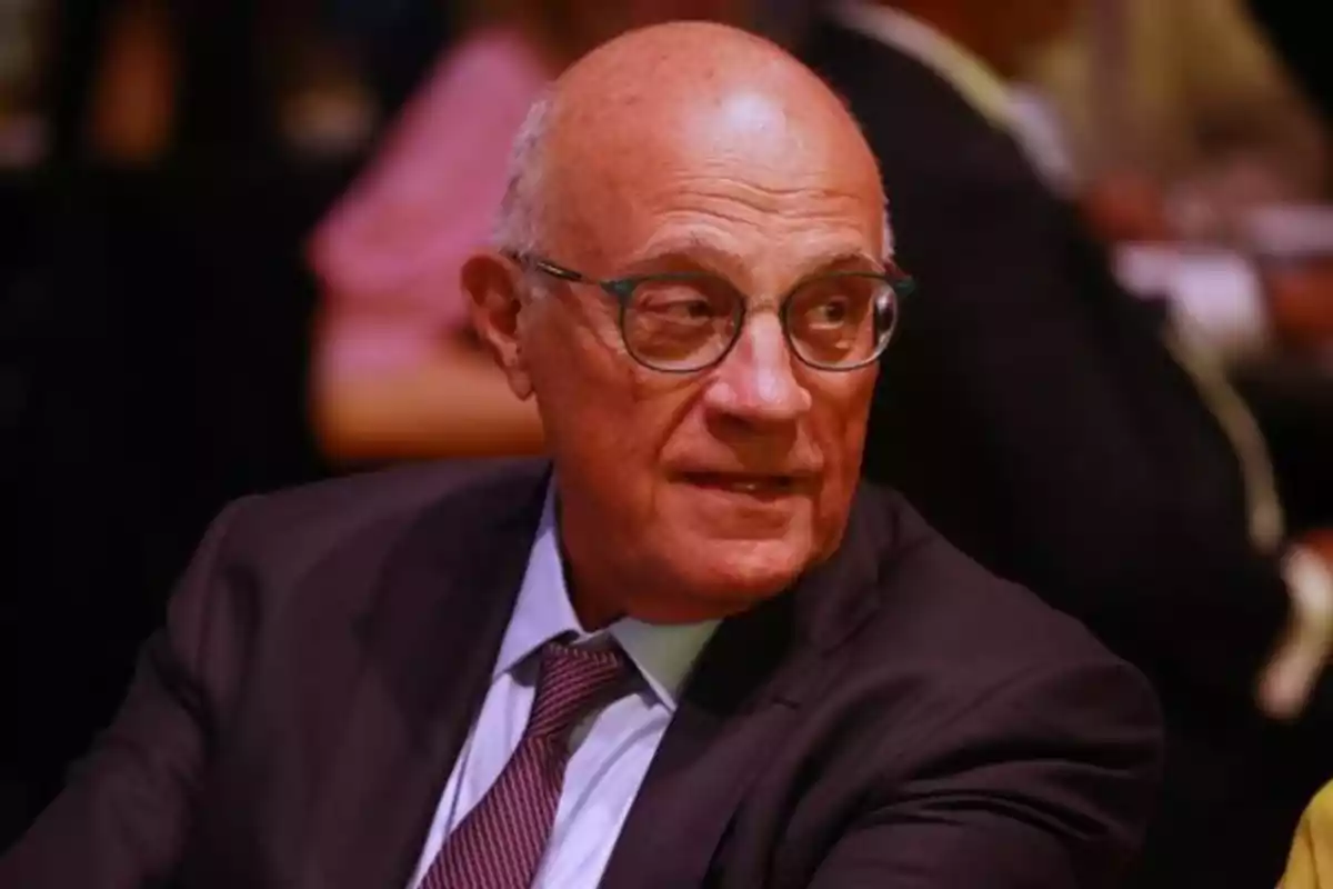 Hombre mayor con gafas y traje oscuro sentado en un evento social mirando hacia un lado