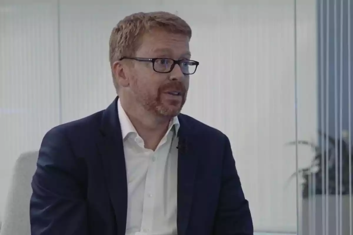 Hombre de cabello corto y barba pelirroja con gafas y traje azul sentado en una oficina moderna Hombre de cabello corto y barba pelirroja con gafas y traje azul sentado en una oficina moderna
