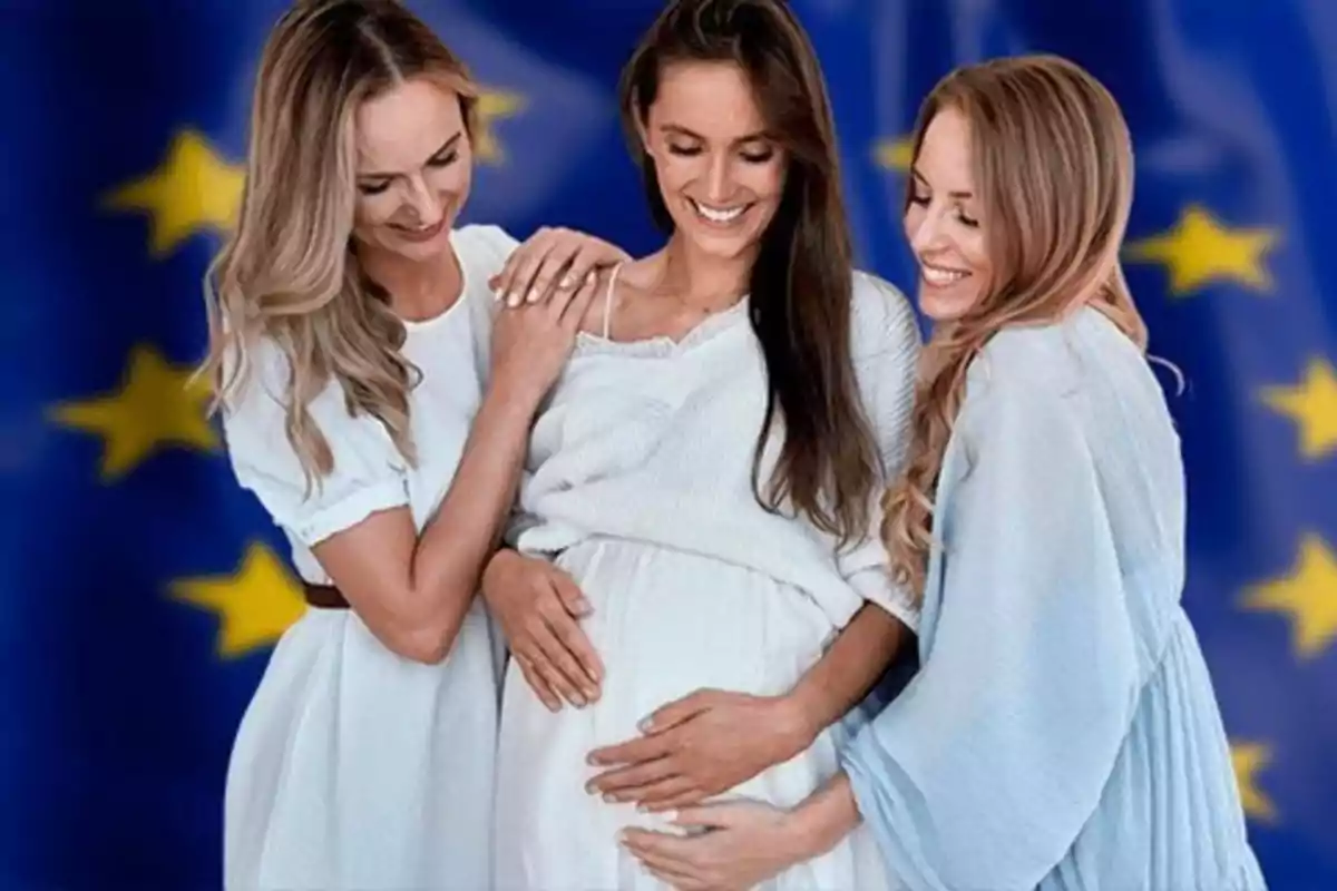 Tres mujeres sonríen mientras una de ellas, embarazada, es abrazada por las otras dos frente a una bandera de la Unión Europea desenfocada.