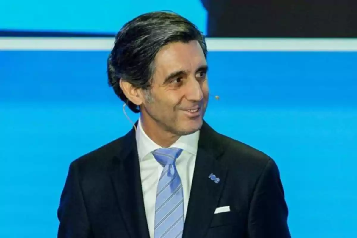 Hombre de cabello canoso con traje oscuro y corbata azul claro hablando en un escenario con fondo azul