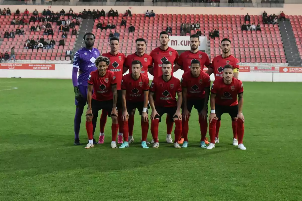 Once jugadores de fútbol posan en el campo antes de un partido con uniforme rojo y negro mientras el portero lleva uniforme morado y en las gradas se ven algunos aficionados Once jugadores de fútbol posan en el campo antes de un partido con uniforme rojo y negro mientras el portero lleva uniforme morado y en las gradas se ven algunos aficionados