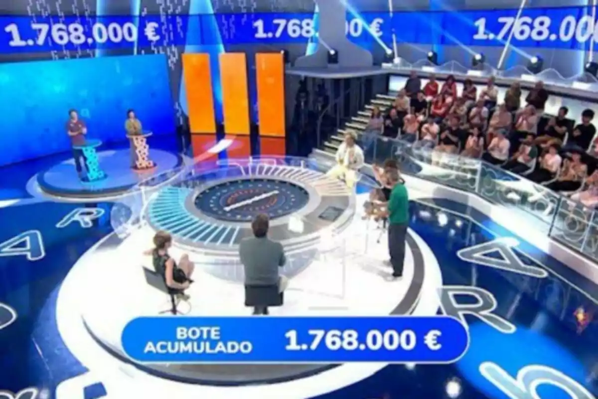 Plano general del plató de 'Pasapalabra' con participantes en un plató moderno, público observando y un premio acumulado de un millón setecientos sesenta y ocho mil euros mostrado en pantalla. Plano general del plató de 'Pasapalabra' con participantes en un plató moderno, público observando y un premio acumulado de un millón setecientos sesenta y ocho mil euros mostrado en pantalla.