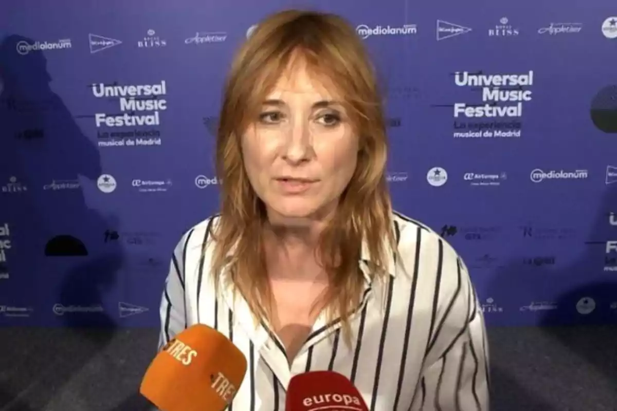 Mujer de cabello rubio rojizo con camisa de rayas blancas y negras hablando frente a varios micrófonos en un evento del Universal Music Festival con fondo azul lleno de logotipos Mujer de cabello rubio rojizo con camisa de rayas blancas y negras hablando frente a varios micrófonos en un evento del Universal Music Festival con fondo azul lleno de logotipos