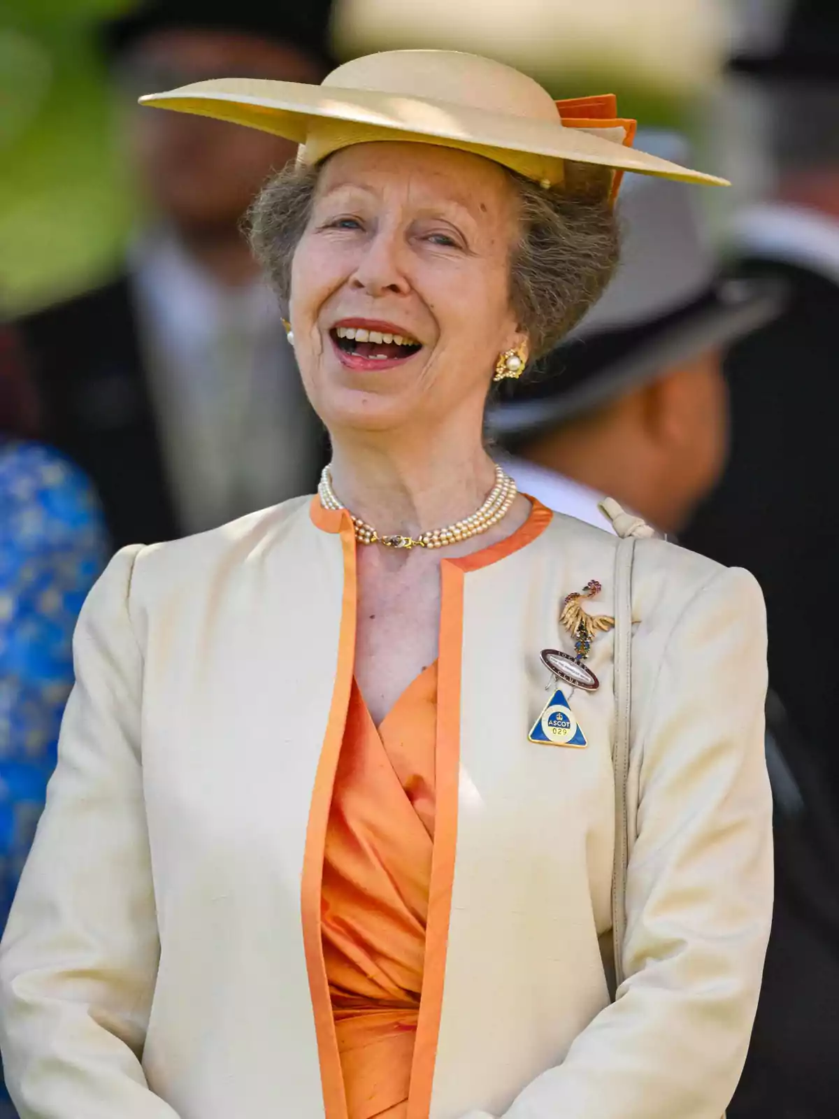 La princesa Ana sonriente con sombrero beige y conjunto elegante en tonos crema y naranja, usando collar de perlas y broches, en un evento al aire libre La princesa Ana sonriente con sombrero beige y conjunto elegante en tonos crema y naranja, usando collar de perlas y broches, en un evento al aire libre