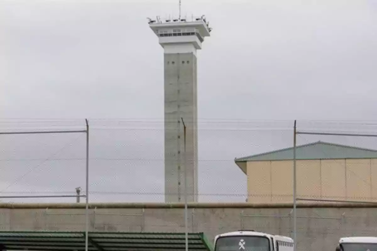 Torre de vigilancia alta en una prisión rodeada de una cerca de alambre y vehículos estacionados al frente Torre de vigilancia alta en una prisión rodeada de una cerca de alambre y vehículos estacionados al frente