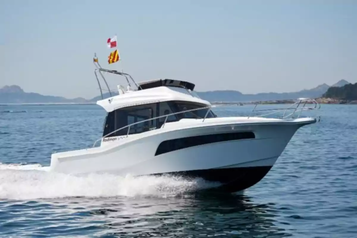 Embarcación blanca navegando a gran velocidad sobre el mar con montañas al fondo y varias banderas ondeando en la parte superior Embarcación blanca navegando a gran velocidad sobre el mar con montañas al fondo y varias banderas ondeando en la parte superior