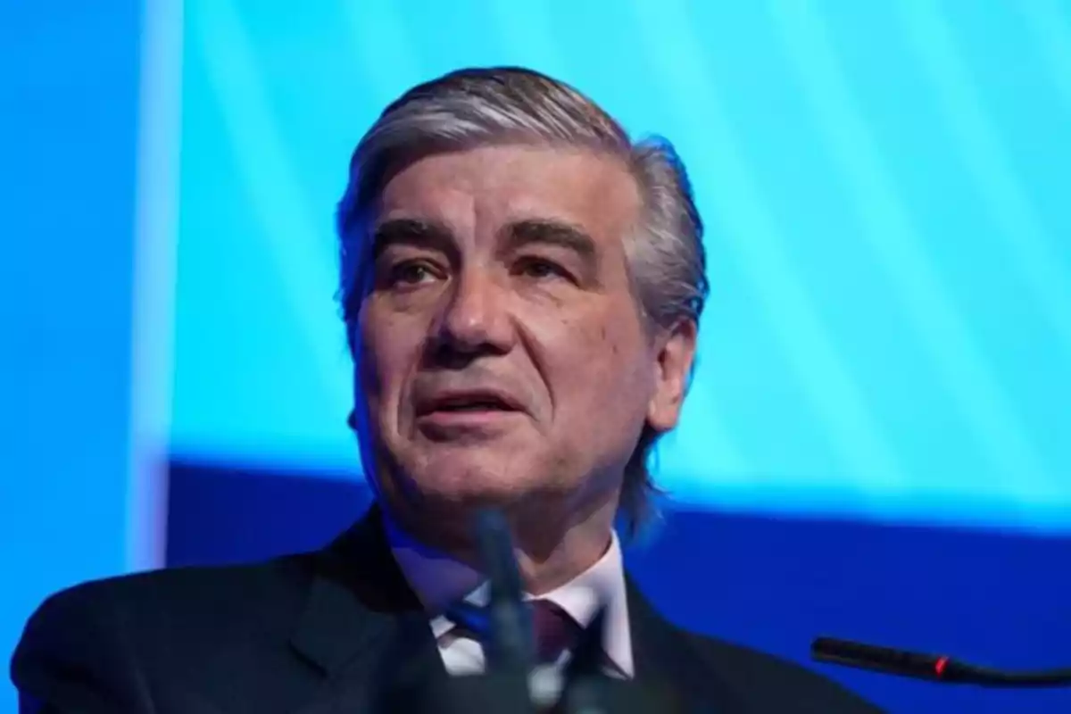 Hombre de cabello canoso y traje oscuro hablando frente a un micrófono con fondo azul